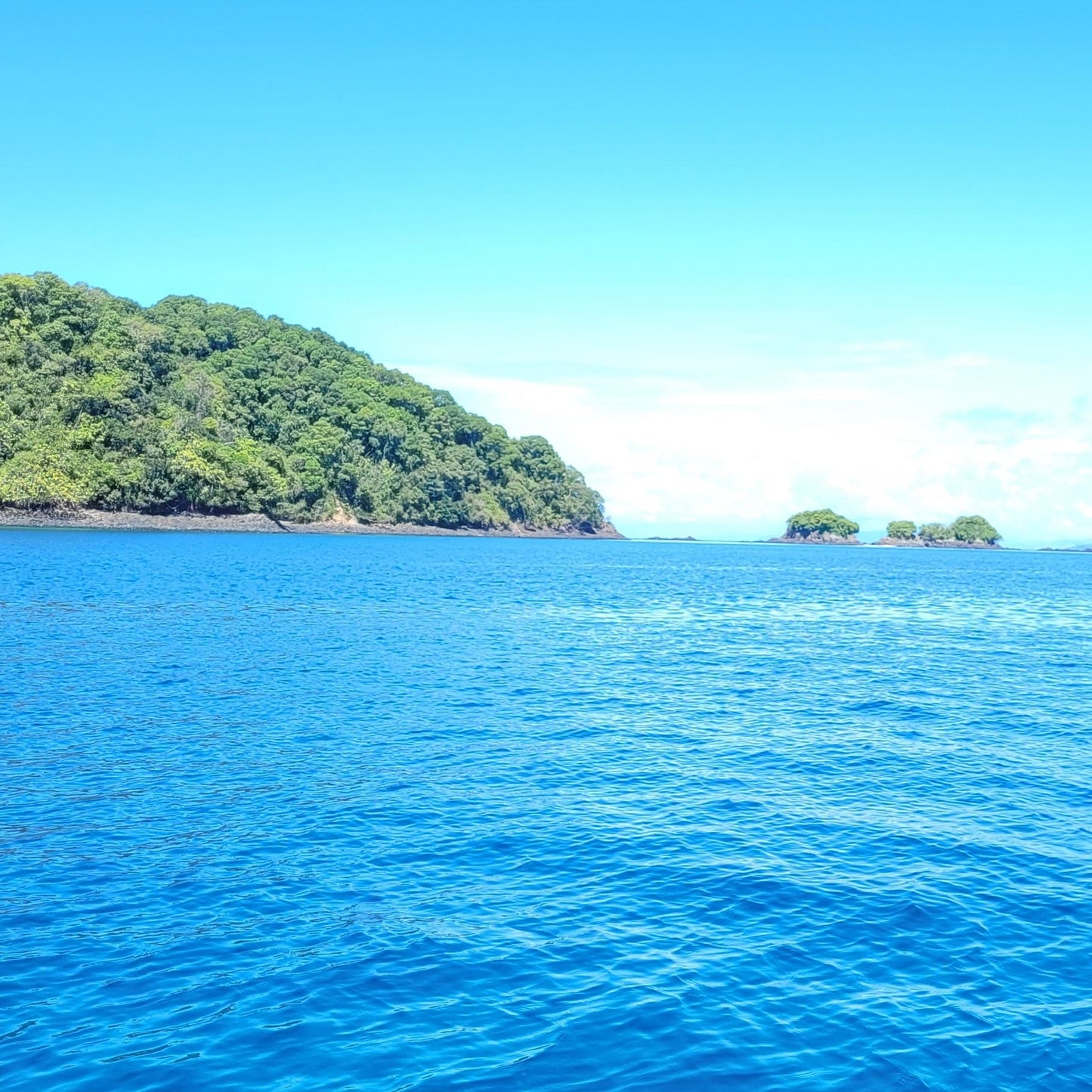 A scenic view of a tropical island with lush green forested hills rising out of deep blue ocean water under a bright blue sky with light clouds.