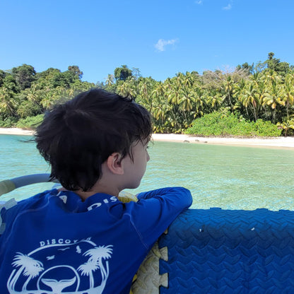 A child wearing a blue t-shirt with a white graphic sits on a boat, looking out at a tropical beach with palm trees and turquoise water under a blue sky.
