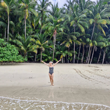 A woman in a dark gray swimsuit stands on a sandy beach with her arms raised, surrounded by tall palm trees and gentle waves in the foreground.