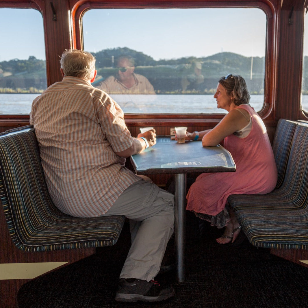 A couple sits at a small table inside a boat or ferry, looking out at water and a shoreline. The man wears a light striped shirt and trousers, and the woman wears a pink dress.