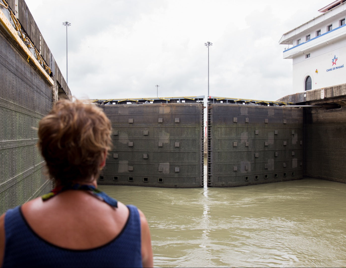 A woman viewed from behind stands in front of a large lock gate structure with murky water below and a white building on the right under an overcast sky.