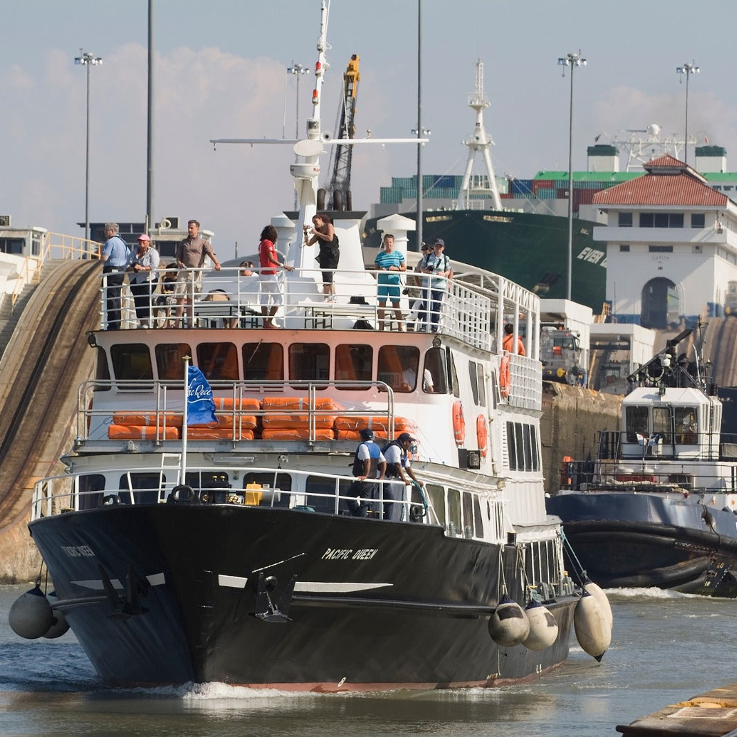 A black and white passenger ferry with orange lifeboats navigating a canal lock, surrounded by other ships and tugboats under a hazy sky.