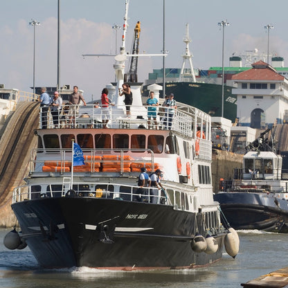 A black and white passenger ferry with orange lifeboats navigating a canal lock, surrounded by other ships and tugboats under a hazy sky.
