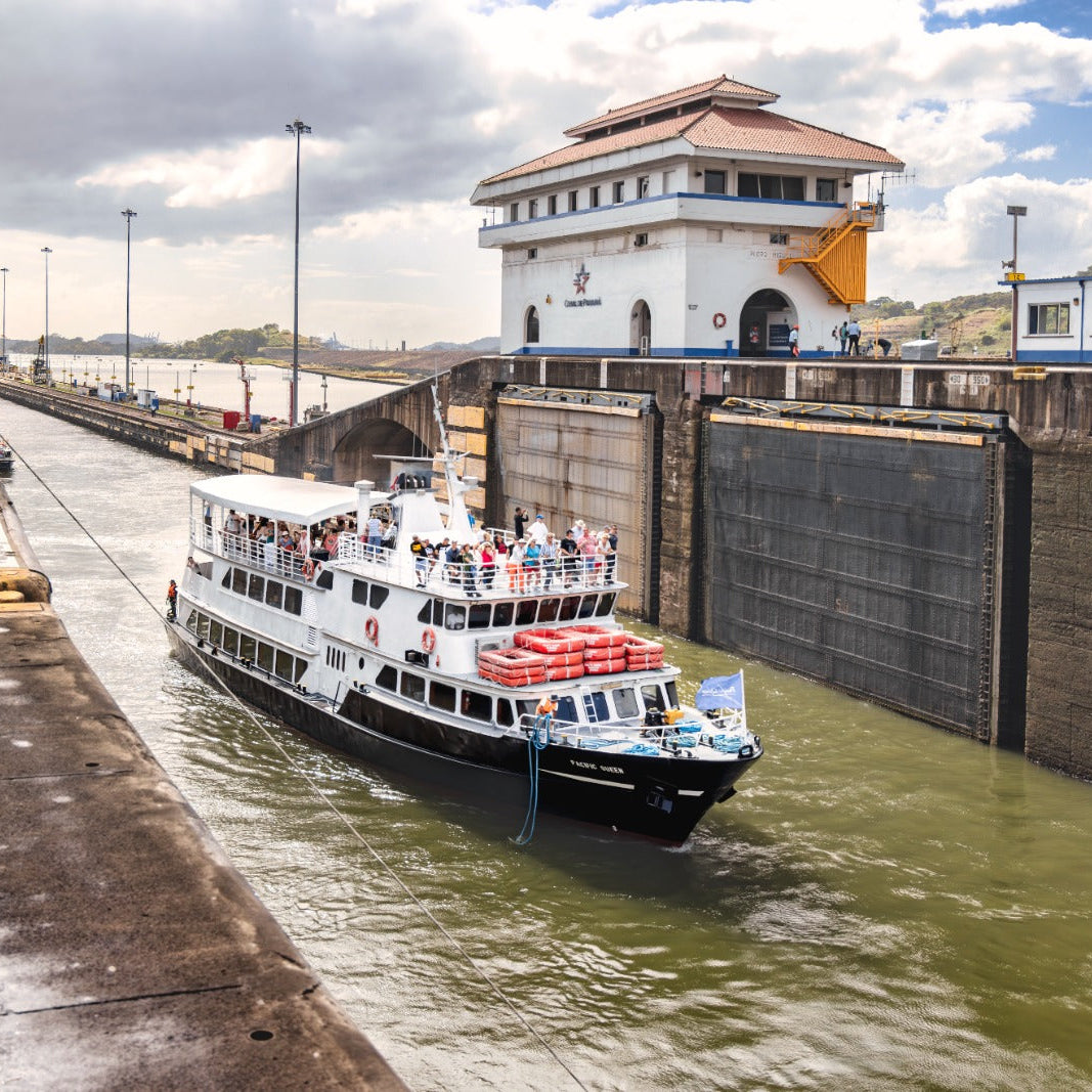 A white passenger ferry with red lifeboats navigates a lock canal next to massive concrete walls under a cloudy sky, with a white and blue control building visible on the right.