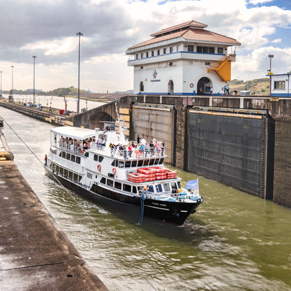 A white passenger ferry with red lifeboats navigates a lock canal next to massive concrete walls under a cloudy sky, with a white and blue control building visible on the right.