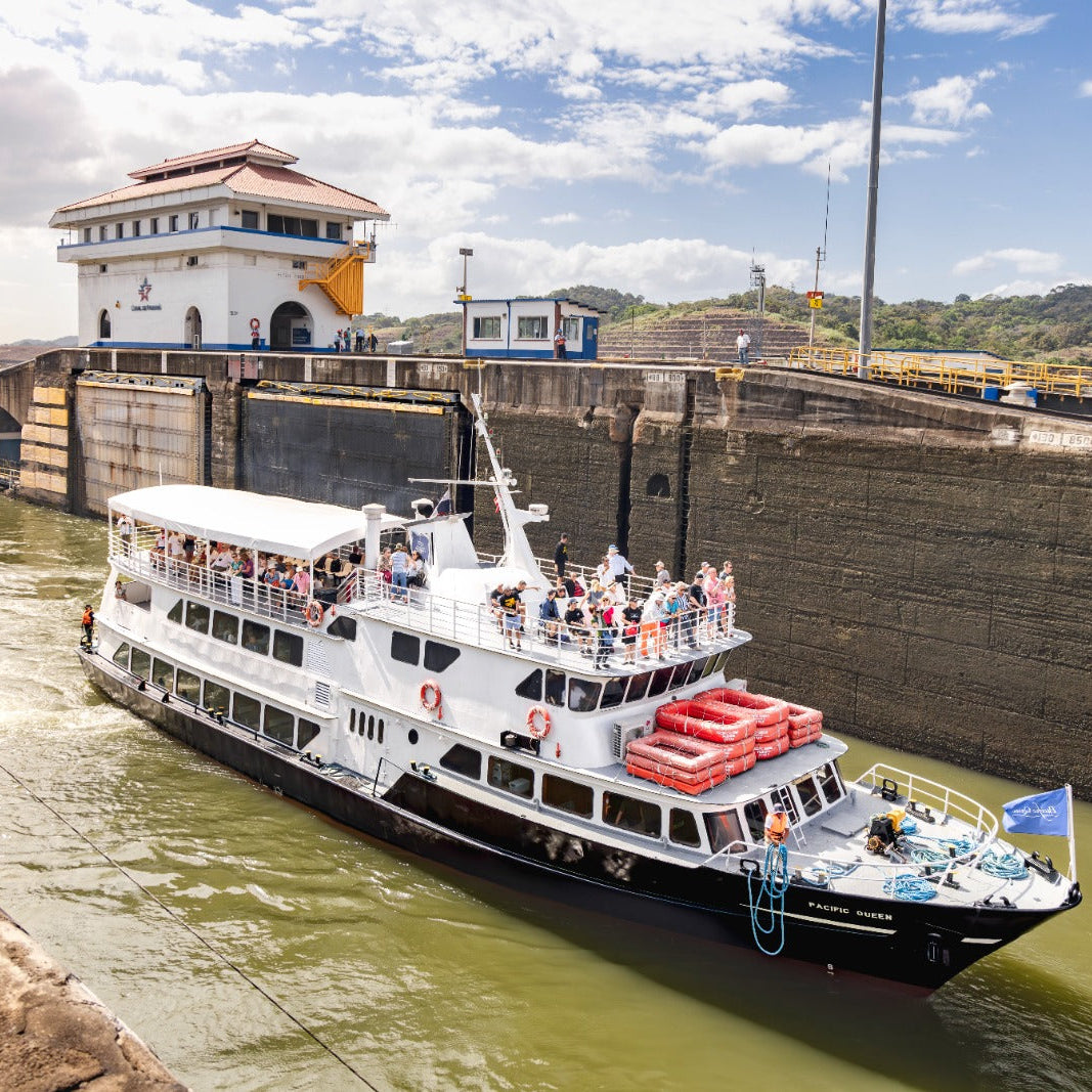A white and black passenger ferry with red lifeboats navigating a lock in a canal under a blue sky with clouds, with a historic building and concrete walls on either side.