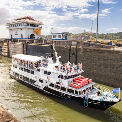 A white and black passenger ferry with red lifeboats navigating a lock in a canal under a blue sky with clouds, with a historic building and concrete walls on either side.