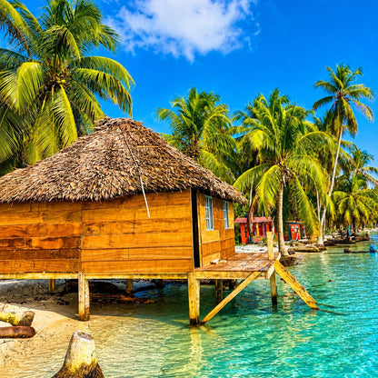 A tropical beach scene with a wooden overwater hut topped with a thatched roof, surrounded by palm trees and clear turquoise water under a bright blue sky.