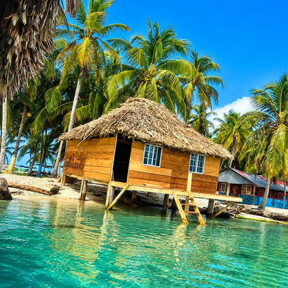 A tropical beach scene with a wooden stilt hut topped with a thatched roof, surrounded by palm trees and clear turquoise water under a bright blue sky.