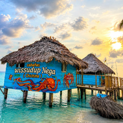 A tropical overwater beach hut with a thatched roof painted bright blue, featuring coral octopus artwork and the text 'cabaPas wissudup Nega (Chichime)'. A second hut and a small thatched lounge area sit on stilts in turquoise water under a dramatic sunset sky with clouds and the sun near the horizon.